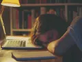 Dziewczyna zasnęła na laptopie, przytłoczona chronicznym zmęczeniem. W tle regały z książkami, lampka nocna.