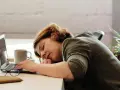 Kobieta z hipersomnia zasnęła przy biurku, opierając głowę na rękach obok laptopa i kubka kawy.