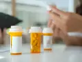 Na stole stoją pomarańczowe aripilek z lekami. W tle widać ręce osoby przeglądającej dokumenty.