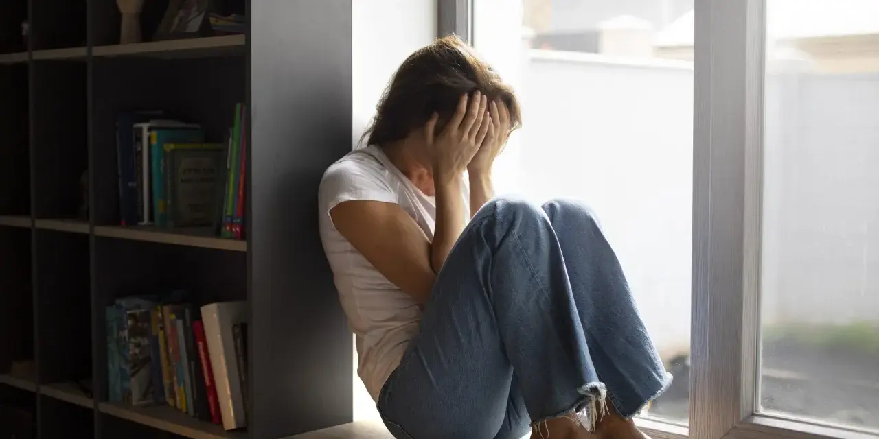 Kobieta siedzi przy oknie, zakrywając twarz dłońmi. Wokół panuje nastrój melancholii, odzwierciedlający dystymię.