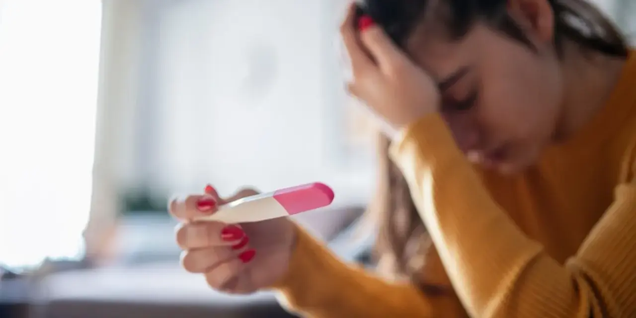 Kobieta z opuszczoną głową trzyma test ciążowy. Jej dłoń z czerwonym lakierem do paznokci zaciska się na teście. Tokofobia może wywoływać takie emocje.