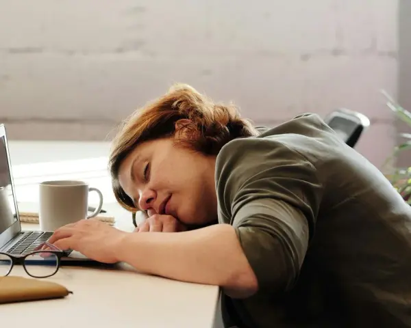 Kobieta z hipersomnia zasnęła przy biurku, opierając głowę na rękach obok laptopa i kubka kawy.