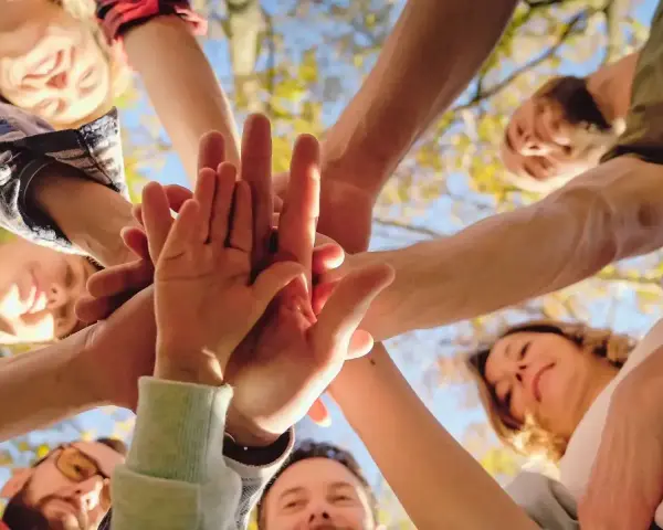 Grupa ludzi złącza dłonie w geście jedności i altruizmu. Uśmiechnięte twarze patrzą w górę, symbolizując wspólny cel i wsparcie.