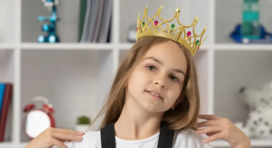 Mała egocentryczka w złotej koronie z klejnotami, pewna siebie, z uśmiechem na twarzy, na tle regału z książkami i zabawkami.