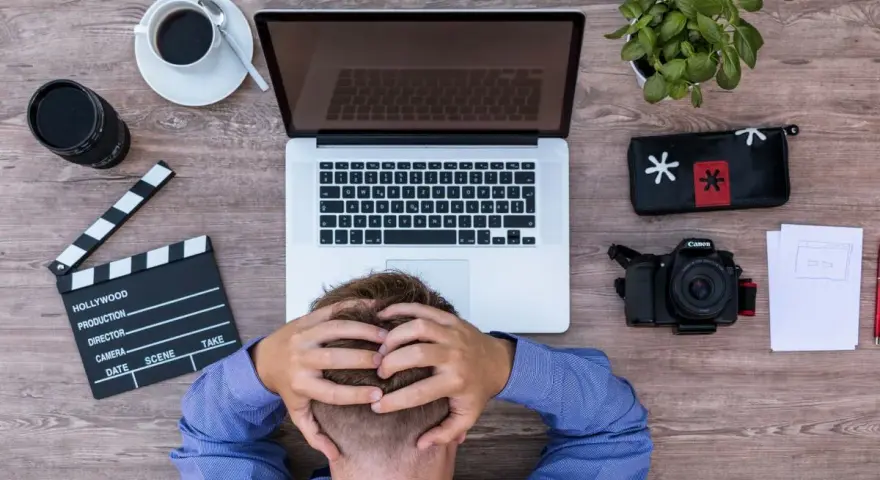Frustracja twórcy filmowego. Na drewnianym biurku leży laptop, aparat, klaps, kawa i roślina.