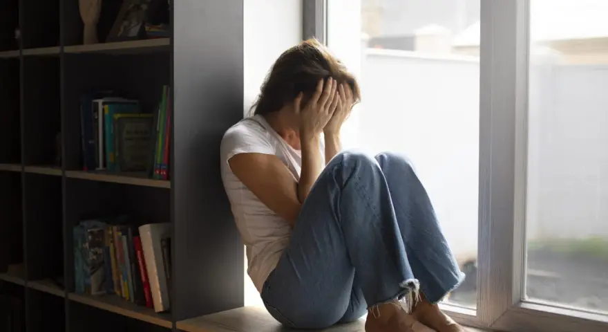 Kobieta siedzi przy oknie, zakrywając twarz dłońmi. Wokół panuje nastrój melancholii, odzwierciedlający dystymię.