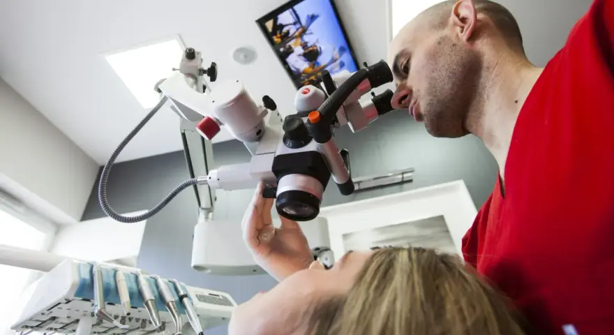 Dentysta w czerwonej bluzie bada pacjentkę przez mikroskop stomatologiczny.