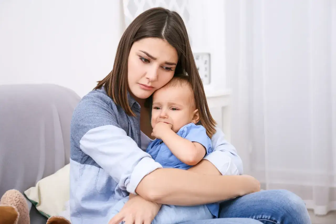 Smutna młoda mama tuli płaczące dziecko. Jej twarz wyraża zmęczenie i troskę, być może to objawy depresji poporodowej.
