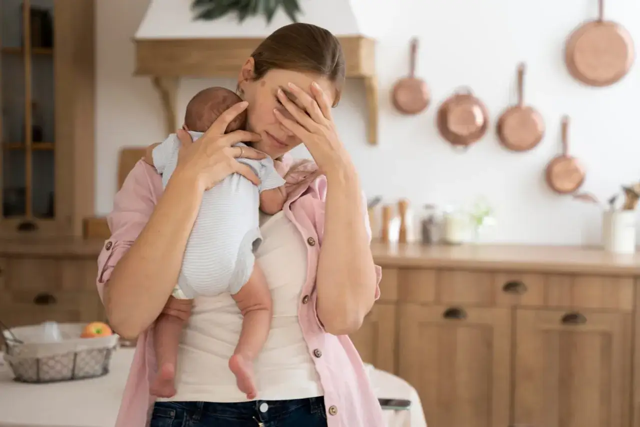 Zmęczona mama z dzieckiem na rękach, czuje baby blues. Dłonią zasłania twarz, wyrażając trudne emocje.