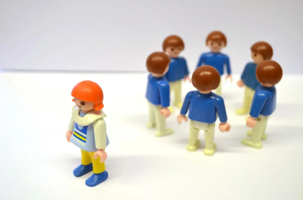 Samotna figurka Playmobil z pomarańczowymi włosami, odwrócona od grupy w niebieskich bluzkach. Scena symbolizuje ostracyzm.