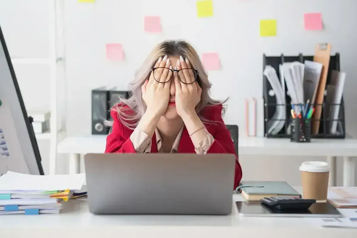 Kobieta w okularach z dłońmi na twarzy, wyrażająca ogromne zmęczenie. Przed nią laptop i stosy dokumentów.