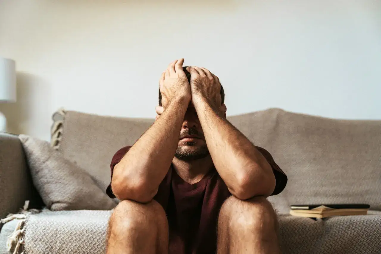 Mężczyzna z rękami na twarzy, siedzący na podłodze przed kanapą. Wygląda na pogrążonego w myślach, być może przeżywa epizod depresyjny. Obok leży otwarta książka.