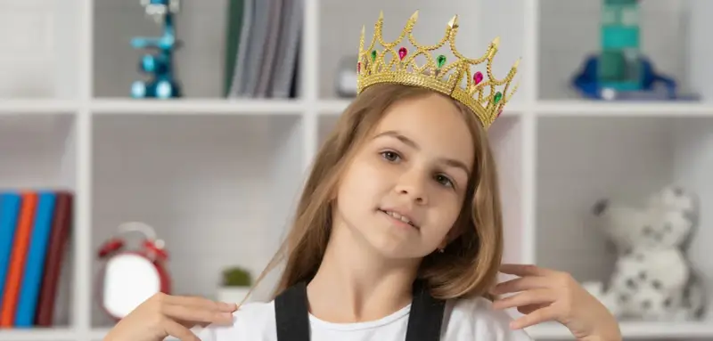 Mała egocentryczka w złotej koronie z klejnotami, pewna siebie, z uśmiechem na twarzy, na tle regału z książkami i zabawkami.