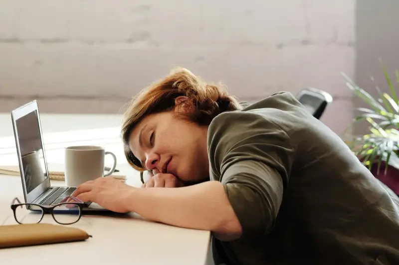 Kobieta z hipersomnia zasnęła przy biurku, opierając głowę na rękach obok laptopa i kubka kawy.